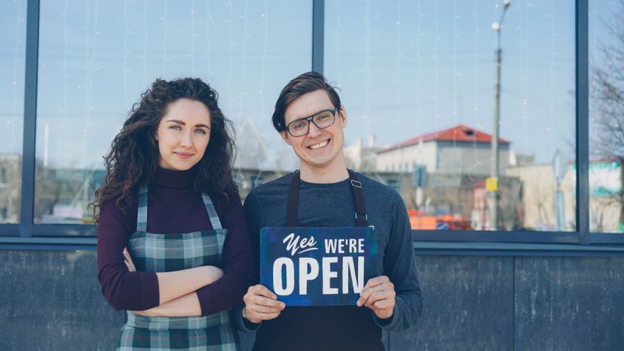 Two small business owners in aprons outside their storefront holding a We’re Open sign