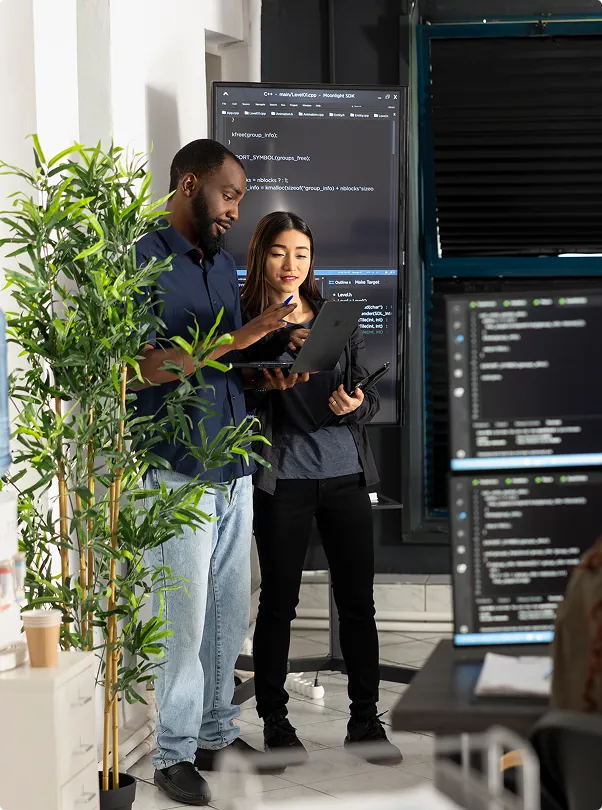 Two developers with a laptop beside monitors full of code